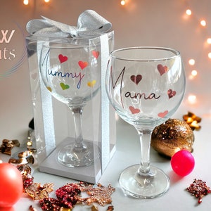 May include: Two clear glass goblets featuring decorative text and heart motifs. One glass is inside a clear plastic box with a silver ribbon, displaying the word Mummy in rainbow colours. The other glass displays the word Mama in rose gold.