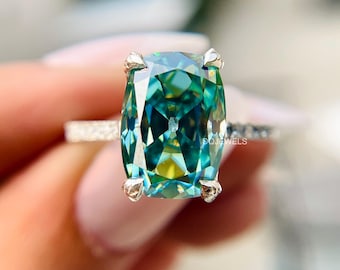 May include: A close-up of a ring featuring a large, rectangular, teal-colored gemstone. The gemstone is set in a silver band with smaller, clear stones. The ring's prongs are silver. The brand name "DQJEWELS" is visible on the ring's band.