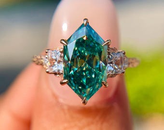 May include: A gold ring featuring a large, emerald-cut, teal-colored gemstone as the centerpiece. Two smaller, square, clear gemstones flank the main stone. The ring has a delicate, ornate band.