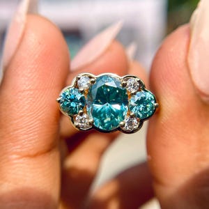 Könnte beinhalten: Ein goldener Ring mit einem ovalen, teal-farbenen Edelstein im Zentrum, flankiert von zwei kleineren teal-farbenen Steinen und akzentuiert mit mehreren kleinen, runden, klaren Edelsteinen. Der Ring wird zwischen zwei Fingern gehalten.