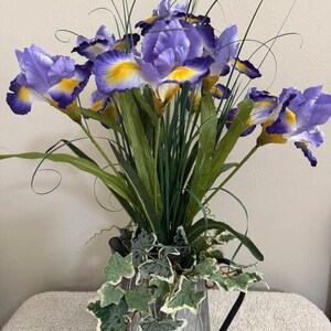 May include: A bouquet of artificial irises in a galvanized metal pitcher. The irises are purple with yellow accents, and the arrangement includes green leaves and decorative ivy. The pitcher has a black handle.