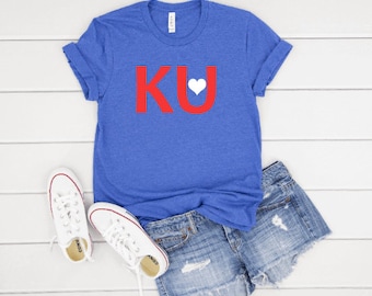 May include: A royal blue t-shirt with the letters "KU" in red with a white heart in the middle of the "U". The shirt is laying on a white wooden surface with a pair of white canvas shoes and blue denim shorts.