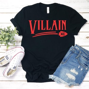 May include: Black t-shirt with red text that says "VILLAIN" with a red arrow pointing to the right and the letters "KC" underneath. The shirt is laying on a wooden surface with a pair of white sneakers and a pair of blue denim shorts.