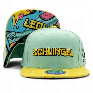 Op de afbeelding: Een lichtgroene snapback-pet met een gele klep en het woord SCHLINGEL in het geel geborduurd op de voorkant. De onderkant van de klep heeft een kleurrijke grafische print met pizzapunten, watermeloen en de tekst LEO.