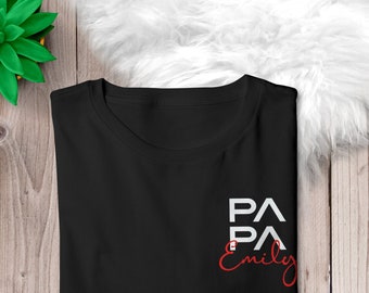 Puede incluir: Camiseta negra con el texto "PAPA" superpuesto a "Emily" en escritura roja. La camiseta está doblada sobre una superficie de madera, con una planta verde y material blanco y esponjoso en el fondo.