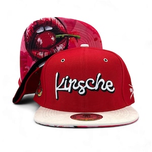 Può includere: Un cappellino da baseball rosso con visiera piatta e la parola Kirsche ricamata in corsivo bianco sul davanti. La parte inferiore della visiera presenta una stampa grafica di labbra rosse che mordono una ciliegia. Un adesivo è centrato.