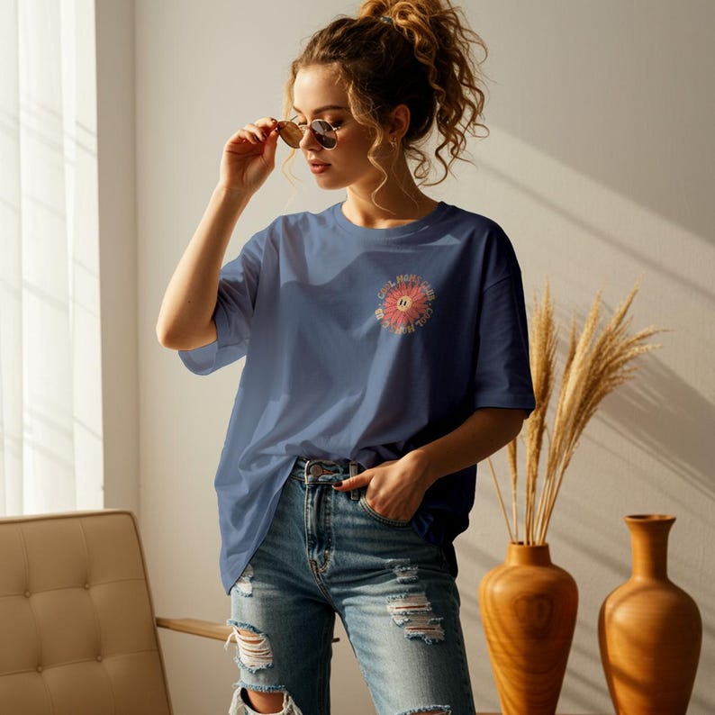 May include: A woman wearing a blue oversized t-shirt with a flower graphic and the text "Get More Sun, Worry Less" in a circle around the flower. She is wearing blue jeans and has her hair pulled back in a ponytail.