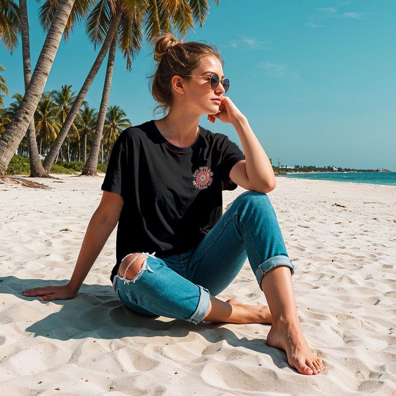 May include: A woman wearing a black t-shirt with a pink flower design and blue jeans sits on a white sandy beach. She is wearing sunglasses and has her hair pulled back in a bun.