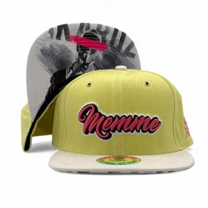Op de afbeelding: Een gele en witte baseballpet met het woord "Memme" in roze en zwarte letters.