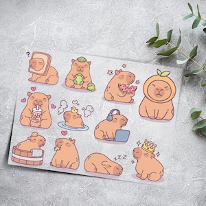 Könnte beinhalten: Ein Stickerbogen mit Cartoon-Wasserschweinen in verschiedenen Posen. Ein Wasserschwein trägt eine Krone, ein anderes isst Wassermelone, und andere sind in einem Bad oder tragen Kopfhörer. Die Aufkleber sind in Braun- und Orangetönen gehalten.
