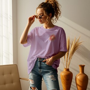May include: A lavender oversized t-shirt with a pink and orange flower graphic on the left chest. The flower has a smiley face in the center.
