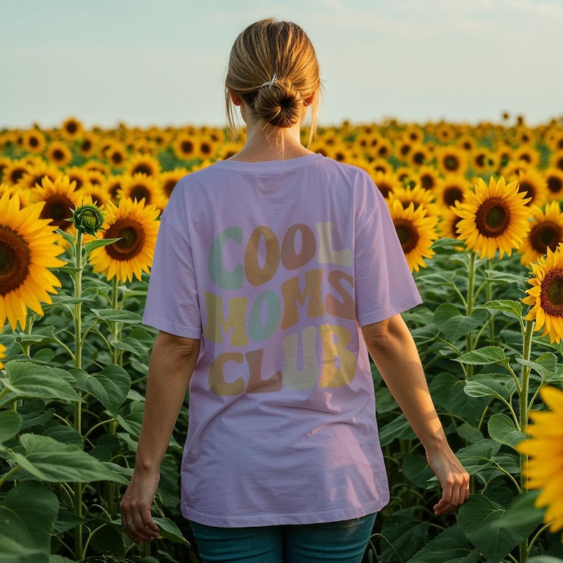 May include: A woman wearing a lavender t-shirt with the text "COOL MOMS CLUB" in colorful, retro-style lettering. She is standing in a field of sunflowers.