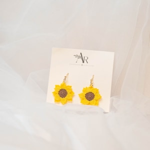 May include: A pair of sunflower earrings with gold hooks. The sunflowers are crocheted with yellow petals and brown centers.