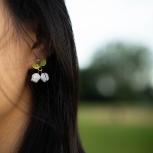 May include: A pair of delicate earrings featuring small white and green crocheted flowers. The earrings are made with a gold-toned metal and have a simple, elegant design.
