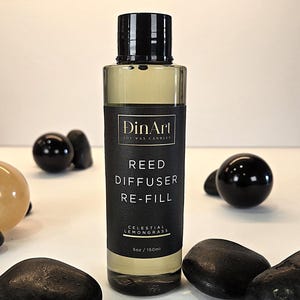 Pode incluir: Uma garrafa de vidro transparente com tampa preta, rotulada como "DinArt Soy Wax Candles" e "Reed Diffuser Re-Fill Celestial Lemongrass". A garrafa cont&eacute;m um l&iacute;quido amarelo e tem 150 ml. Pedras e esferas decorativas pretas circundam a garrafa.