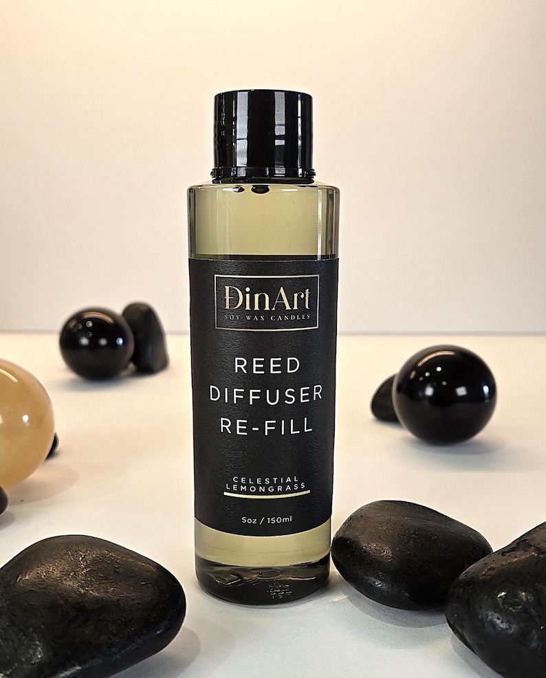 Pode incluir: Uma garrafa de vidro transparente com tampa preta, rotulada como "DinArt Soy Wax Candles" e "Reed Diffuser Re-Fill Celestial Lemongrass". A garrafa cont&eacute;m um l&iacute;quido amarelo e tem 150 ml. Pedras e esferas decorativas pretas circundam a garrafa.