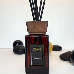 Peut inclure: Un diffuseur en verre ambré foncé avec un bouchon en bois et des roseaux noirs. L'étiquette indique "PinArt Candles Celestial Lemongrass 150 ml / 5 oz". Des pierres noires et une sphère jaune sont en arrière-plan.