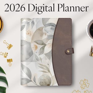 Pode incluir: Um planejador digital de 2026 com um design floral em aquarela em tons neutros. O planejador tem uma capa de couro marrom com fecho de botão. Clipes de papel dourados, uma vela e uma xícara de café também são visíveis.