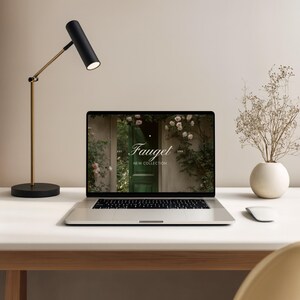 May include: A modern desk setup featuring a laptop displaying the "Fauget New Collection" website. A black and gold desk lamp illuminates the scene, complemented by a white vase with dried flowers and a wireless mouse. The desk is white, and the background is a neutral beige.
