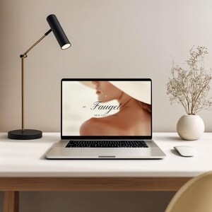 May include: A modern desk setup featuring a laptop displaying a website with the text "Fauget New Collection." A black and gold desk lamp illuminates the laptop. A small white vase with dried flowers and a white mouse complete the scene.