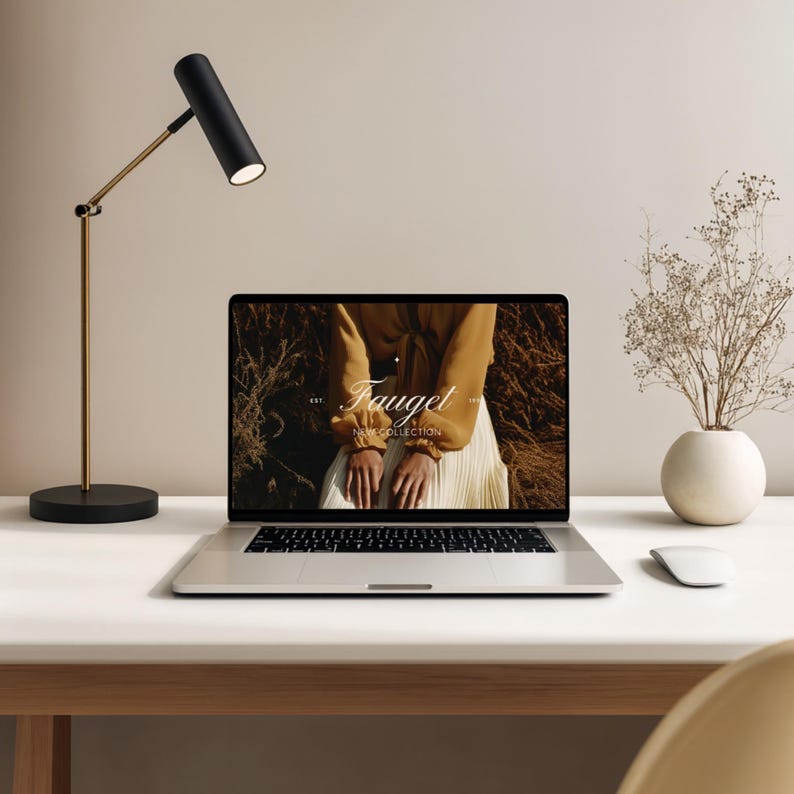 May include: A modern desk setup featuring a laptop displaying a website with the text "Fauget". A black and gold desk lamp illuminates the scene. A small white vase with dried flowers and a wireless mouse complete the minimalist aesthetic.