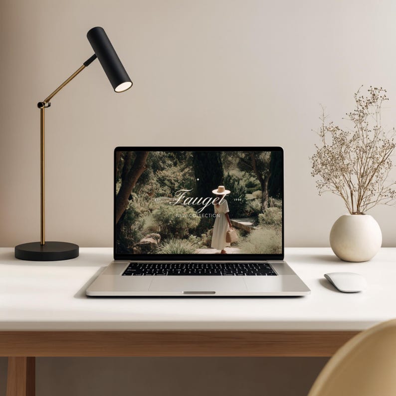 May include: A modern desk setup featuring a laptop displaying a website with the word "Fauget". A black and gold desk lamp is positioned to the left. A white vase with dried flowers and a computer mouse are also on the desk.