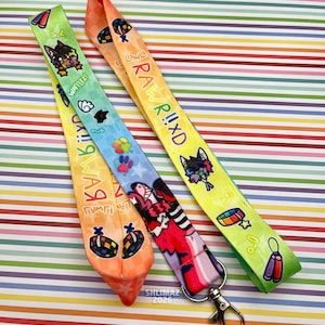 May include: Three colorful lanyards with cartoon graphics and text. The lanyards feature a variety of designs, including cartoon animals, paw prints, and text that reads "RAWRI!XD" and "WaffleZ!". Each lanyard has a metal clasp.