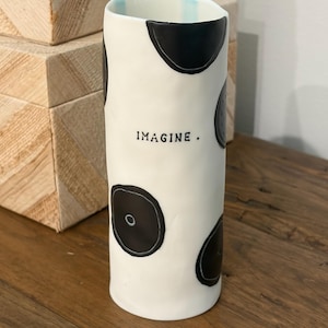 Puede incluir: Un jarrón de cerámica blanco con diseños circulares negros y la palabra "IMAGINE" en negro. El jarrón tiene un borde superior ligeramente irregular y un borde interior azul claro. El jarrón está sobre una superficie de madera.