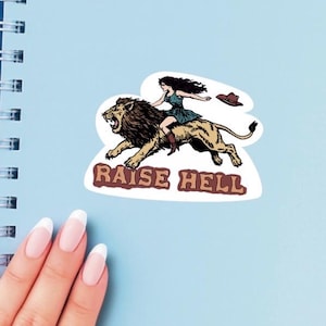 May include: A light blue spiral notebook with a sticker featuring a woman riding a lion, with the text "RAISE HELL". The notebook is on top of a dark blue notebook. The image is taken from a top-down perspective.