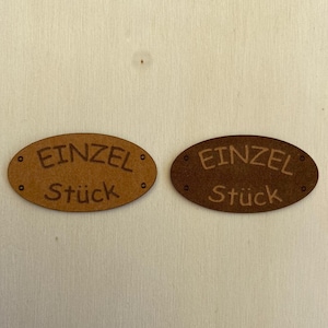 Puede incluir: Dos letreros ovalados de madera marrón con el texto "Einzel Stück" en letras blancas.