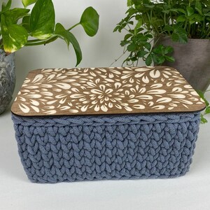 Könnte beinhalten: Eine blaue gestrickte Aufbewahrungsbox mit einem Holzdeckel mit einem weißen Blumenmuster. Die Box steht auf einer weißen Oberfläche.