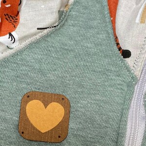 Puede incluir: Una prenda de vestir de tela verde con un parche de corazón marrón cosido en la parte delantera. El parche tiene un corazón amarillo en el interior.