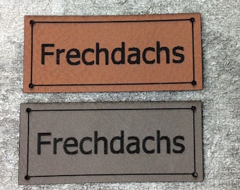 May include: Two leather labels with the text "Frechdachs" printed in black. The top label is brown and the bottom label is grey.