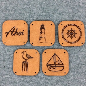 Puede incluir: Cinco etiquetas cuadradas de madera con temas náuticos. Las etiquetas presentan un faro, un velero, una gaviota, un timón de barco y la palabra "¡Ahoy!"