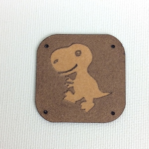 Puede incluir: Parche de cuero marrón con un gráfico de dinosaurio marrón claro. El parche tiene cuatro agujeros alrededor de los bordes.