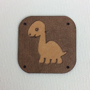 Op de afbeelding: Een bruin leren patch met een uitsnijding van een lichtbruine dinosaurus. De patch heeft vier zwarte stikgaatjes rond de randen.