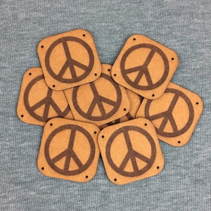 Puede incluir: Un grupo de siete parches de cuero marrón con un diseño de signo de paz. Los parches están dispuestos en un círculo suelto sobre un fondo de tela gris.