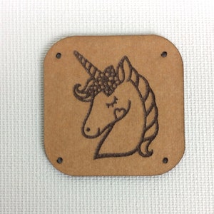 Puede incluir: Un cuadrado de madera marrón con esquinas redondeadas que presenta un diseño de unicornio con los ojos cerrados cortado con láser. El unicornio tiene un cuerno, una melena y una cola. Hay cuatro agujeros alrededor de los bordes del cuadrado.