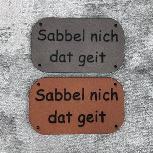 Op de afbeelding: Twee rechthoekige leren patches met de tekst "Sabbel nich dat geit" in zwarte letters. De bovenste patch is grijs en de onderste patch is bruin.
