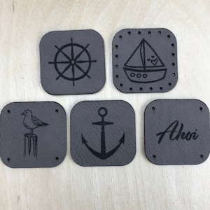 Può includere: Cinque toppe in pelle grigia con disegni nautici. Le toppe presentano un timone, una barca a vela, un gabbiano, un'ancora e la parola "Ahoy".