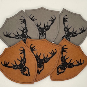 May include: A selection of shield-shaped leather patches in grey and brown. Each patch features a black silhouette of a stag's head with antlers. The patches are arranged in a layered pattern, with a textured finish.
