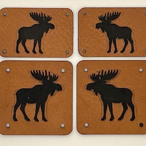 以下が含まれることがあります： 黒いヘラジカのシルエットデザインが施された、正方形の茶色の革製コースター4枚。各コースターにはヘラジカの画像があり、各コーナーに小さなリベットで固定されています。コースターは2x2のグリッドに配置されています。