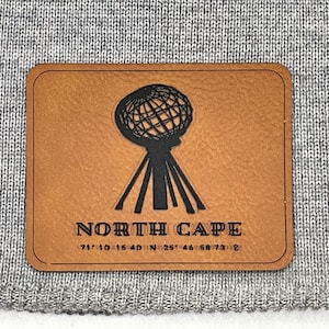 Op de afbeelding: Een grijze gebreide beanie met een bruin leren patch. De patch toont een zwart wereldbolontwerp boven de woorden "NORTH CAPE" en coördinaten. De beanie is gemaakt van een zacht, getextureerd materiaal.