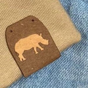 Puede incluir: Dos parches de cuero marrón con una silueta de rinoceronte en cada uno. Los parches están cosidos en una tela vaquera azul claro.