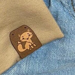 Può includere: Due toppe in pelle marrone con un design di volpe dei cartoni animati cucite su un paio di jeans blu chiaro.