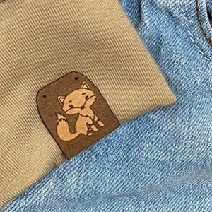 May include: A pair of blue denim jeans with a brown leather patch featuring a cartoon fox on the right pocket. The jeans have a tan fabric cuff on the left leg.