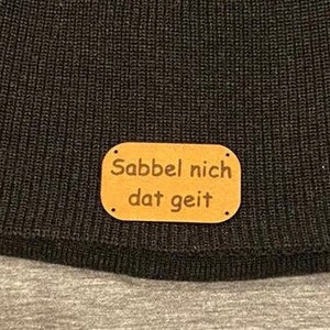 Peut inclure: Bonnet en tricot marron foncé avec un petit patch rectangulaire en cuir beige. Le patch porte l'inscription "Sabbel nich dat geit" en noir. Le bonnet est en tricot côtelé doux.