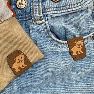 Könnte beinhalten: Blaue Jeans aus Denim mit zwei braunen Lederflicken, die ein Cartoon-Bärenmotiv zeigen. Die Flicken sind auf der Vorderseite der Jeans angenäht.