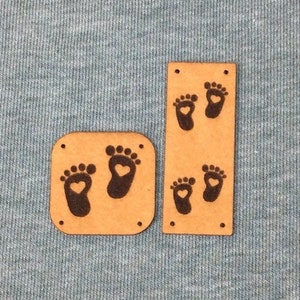 Puede incluir: Dos recortes de madera con forma de pies de bebé con detalles de corazón. Uno es cuadrado, el otro rectangular. Los pies del bebé son negros y la madera es de color marrón claro. Sobre un fondo de tela azul claro.