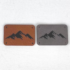 Può includere: Due toppe rettangolari con un disegno di catena montuosa. Una è marrone con una grafica di montagna nera, e l'altra è grigia con una grafica di montagna nera. Ogni toppa ha quattro piccoli fori negli angoli.